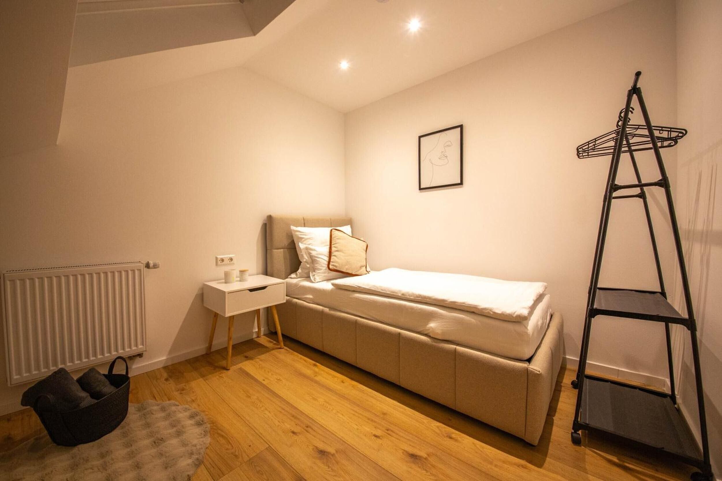 A small, cozy bedroom with a single bed, a bedside table, a radiator, a black metal shelf, and a fluffy rug.
