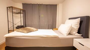 Una cama bien hecha con un cojín, una mesita de noche y una estantería metálica.