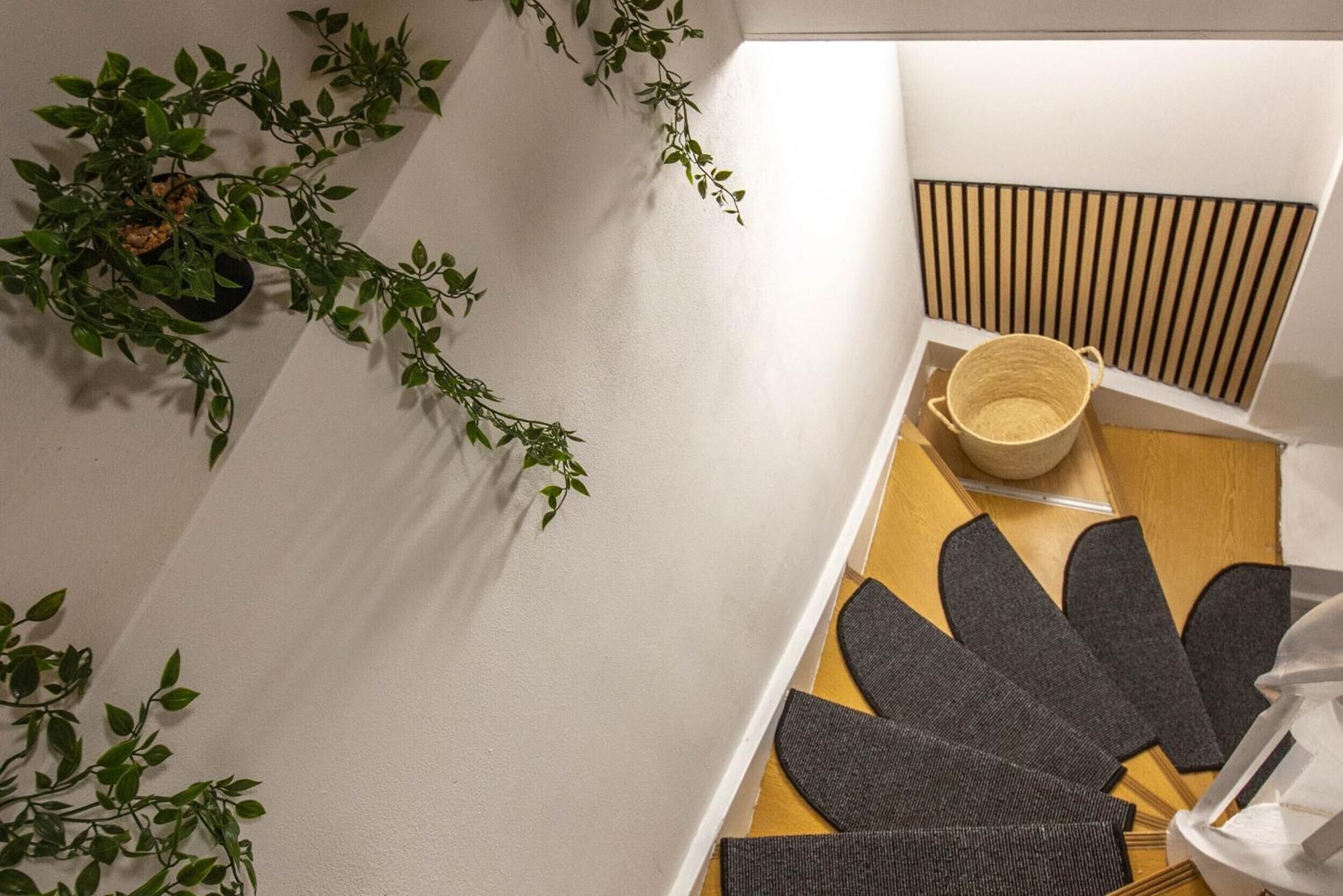 Une pièce avec un mur blanc, un banc en bois à lattes, un panier tressé et une paire de chaussons gris.