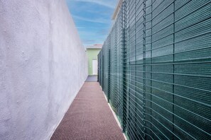 Ein schmaler Gang mit strukturierter Bodenfläche, einem grünen Metallzaun an der Seite und einer weißen Wand.