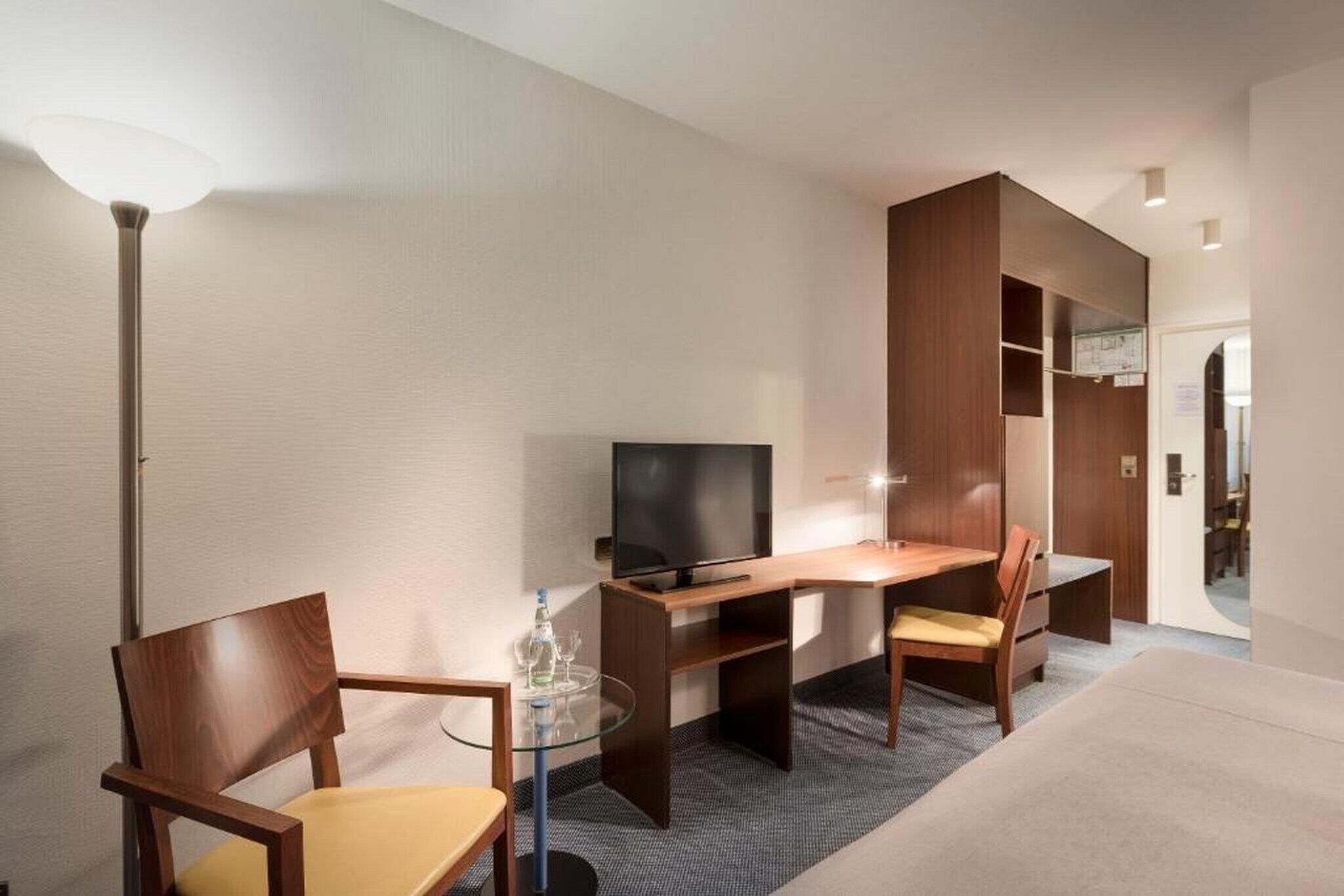 A hotel room with a bed, a desk with a television, two chairs, a glass table, and a wardrobe.