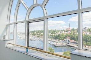 Udsigt fra et vindue med gelænder, der viser en lysthavn med både og en kirketårn i det fjerne.