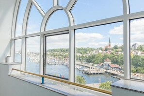 Uitzicht vanuit een raam met een hekwerk, uitkijkend over een jachthaven met boten en een kerktoren in de verte.