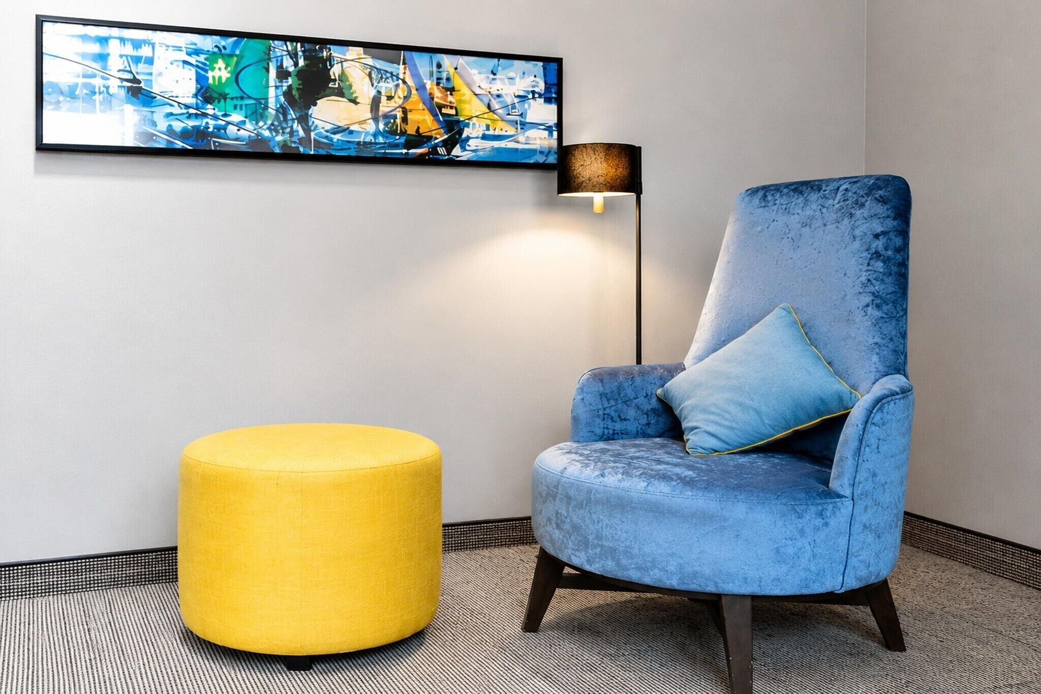 A room with a blue armchair, a yellow ottoman, a floor lamp, and a large abstract painting on the wall.