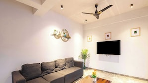A living room with a sectional sofa, a coffee table with a vase of flowers, a ceiling fan, and a flat-screen TV mounted on the wall.