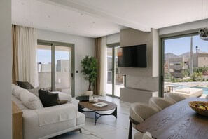 A modern living room with a sofa, coffee table, and dining table, a TV mounted on the wall, and a large window with a view of the outdoors. - Pyxis Villa, Family Friendly with Heated Pool (Perama)