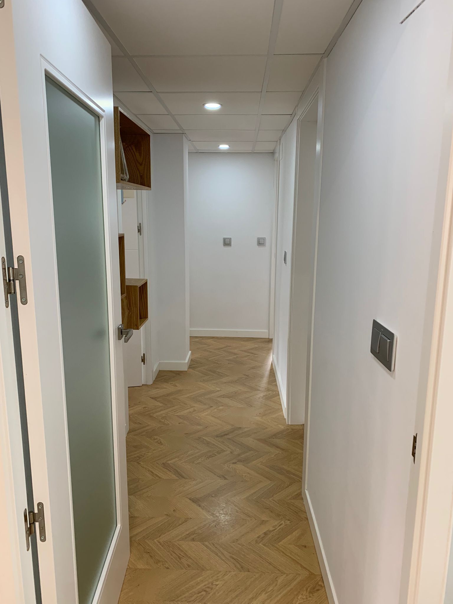 Un couloir doté d’un plancher en bois, de murs blancs et d’une porte avec un panneau en verre dépoli.