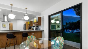 Uma cozinha moderna com uma mesa de jantar de vidro, cadeiras coloridas e vista para um jardim através de uma porta deslizante.
