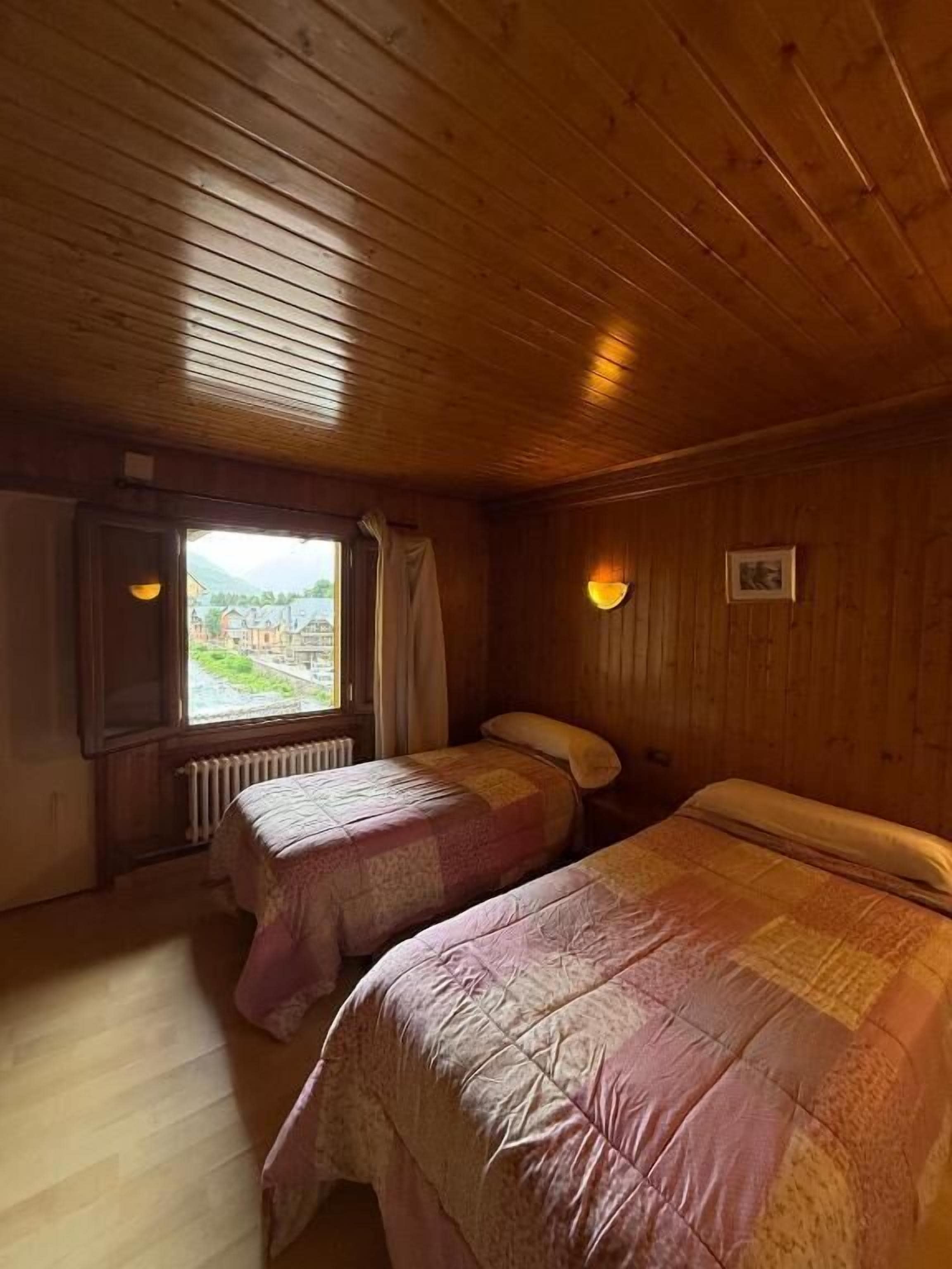 A room with wooden walls and ceiling, two beds with patterned bedding, a window with a view, and a radiator.