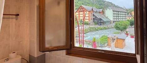 Un baño con techo de madera, una ventana con vistas a edificios y follaje, un inodoro, un bidé y un radiador.