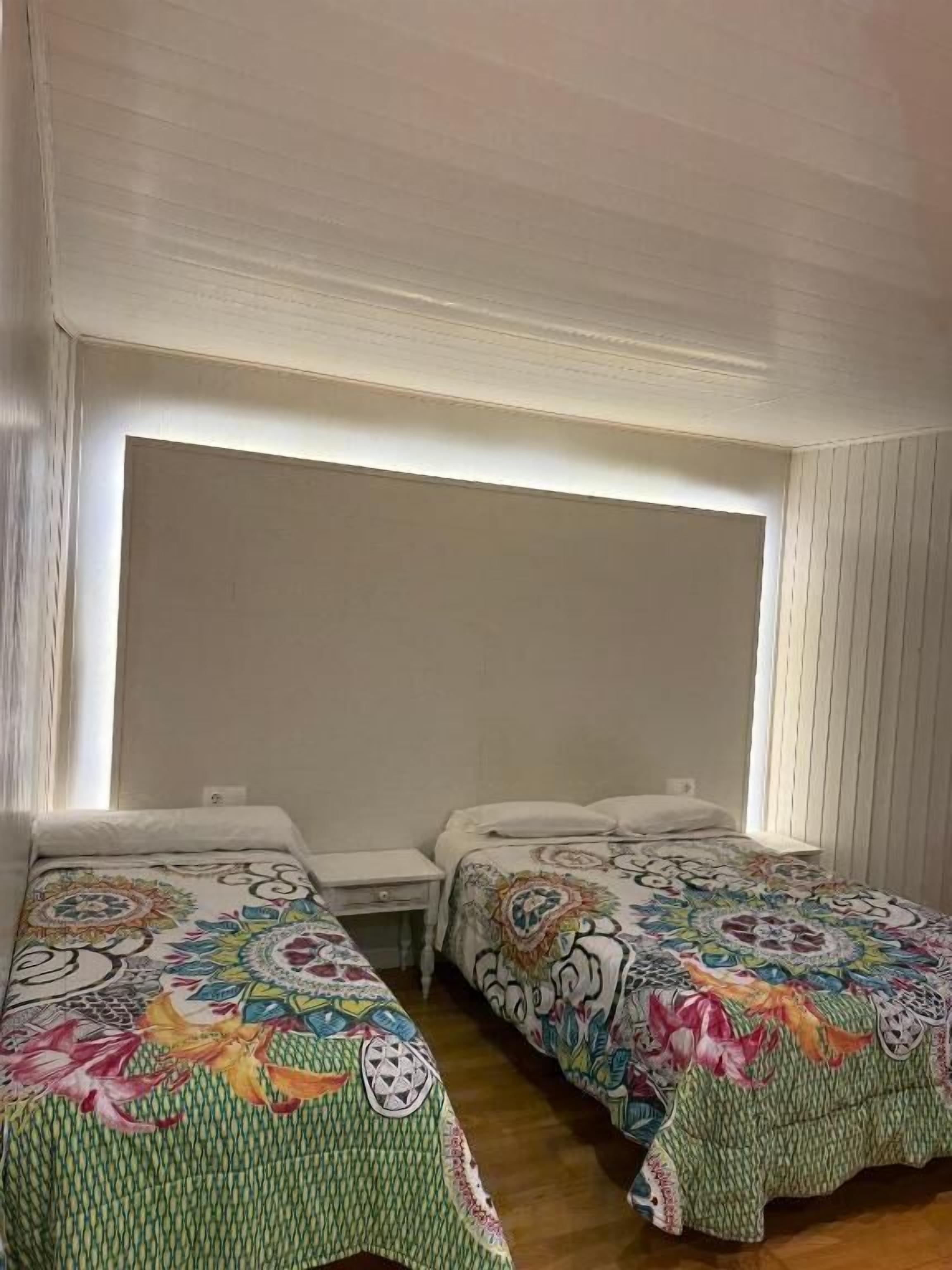 Een kamer met twee bedden, elk voorzien van een kleurrijk sprei, een nachtkastje en een raam met jaloezieën.