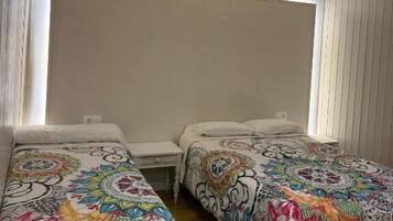 A room with two beds, each with a colorful bedspread, a bedside table, and a window with blinds.