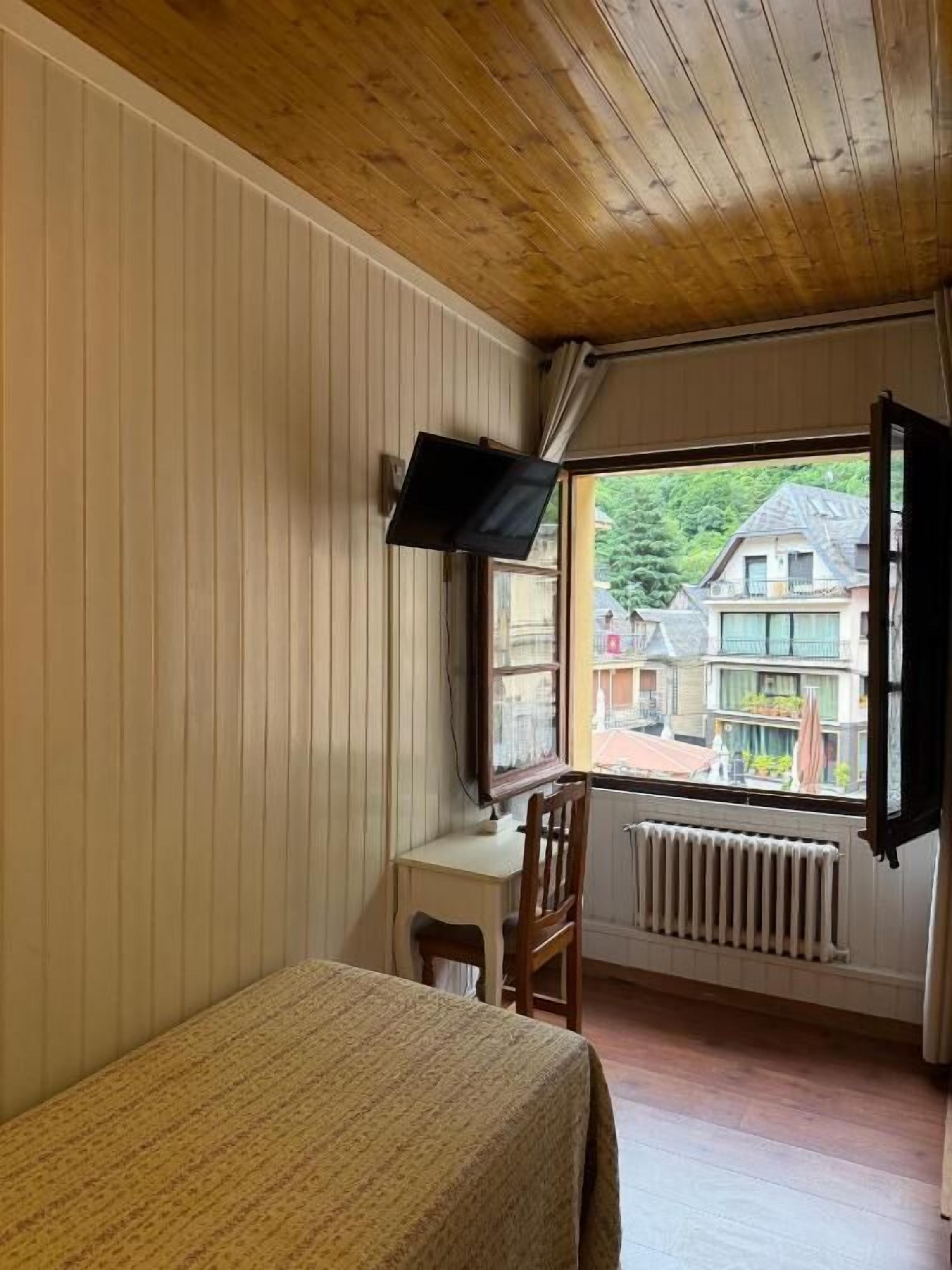 Ein Zimmer mit einem Bett, einem Schreibtisch, einem Sessel, einem Fernseher und einem Fenster mit geöffnetem Vorhang.