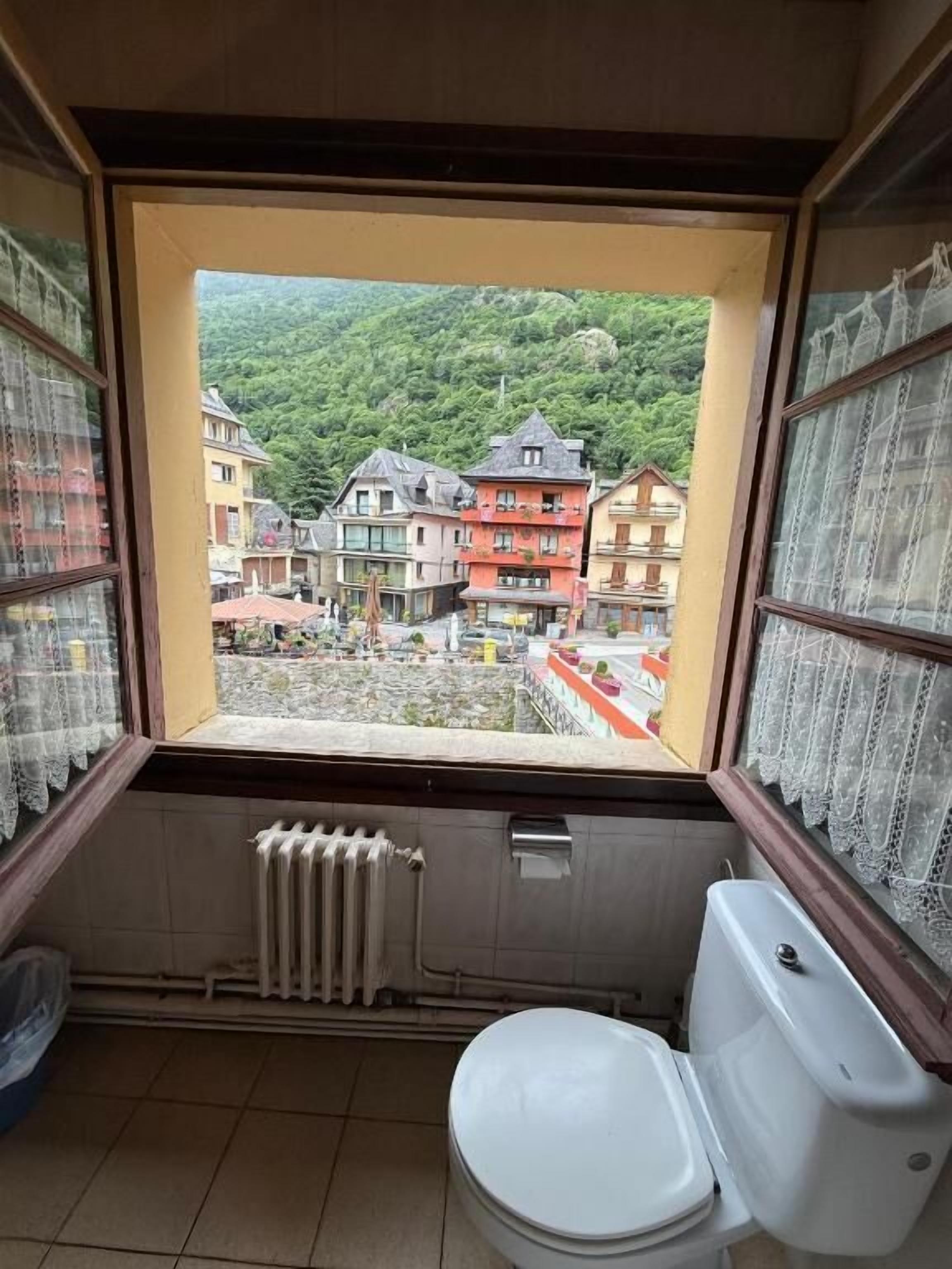 Ein kleines Badezimmer mit Toilette, einem Heizkörper und einem Fenster mit Blick auf eine Hügellandschaft mit Gebäuden.