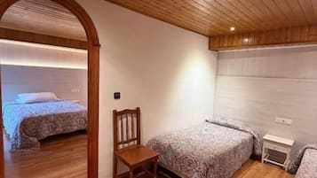 A room with wooden flooring, two beds with patterned bedding, a wooden chair, and a small bedside table.