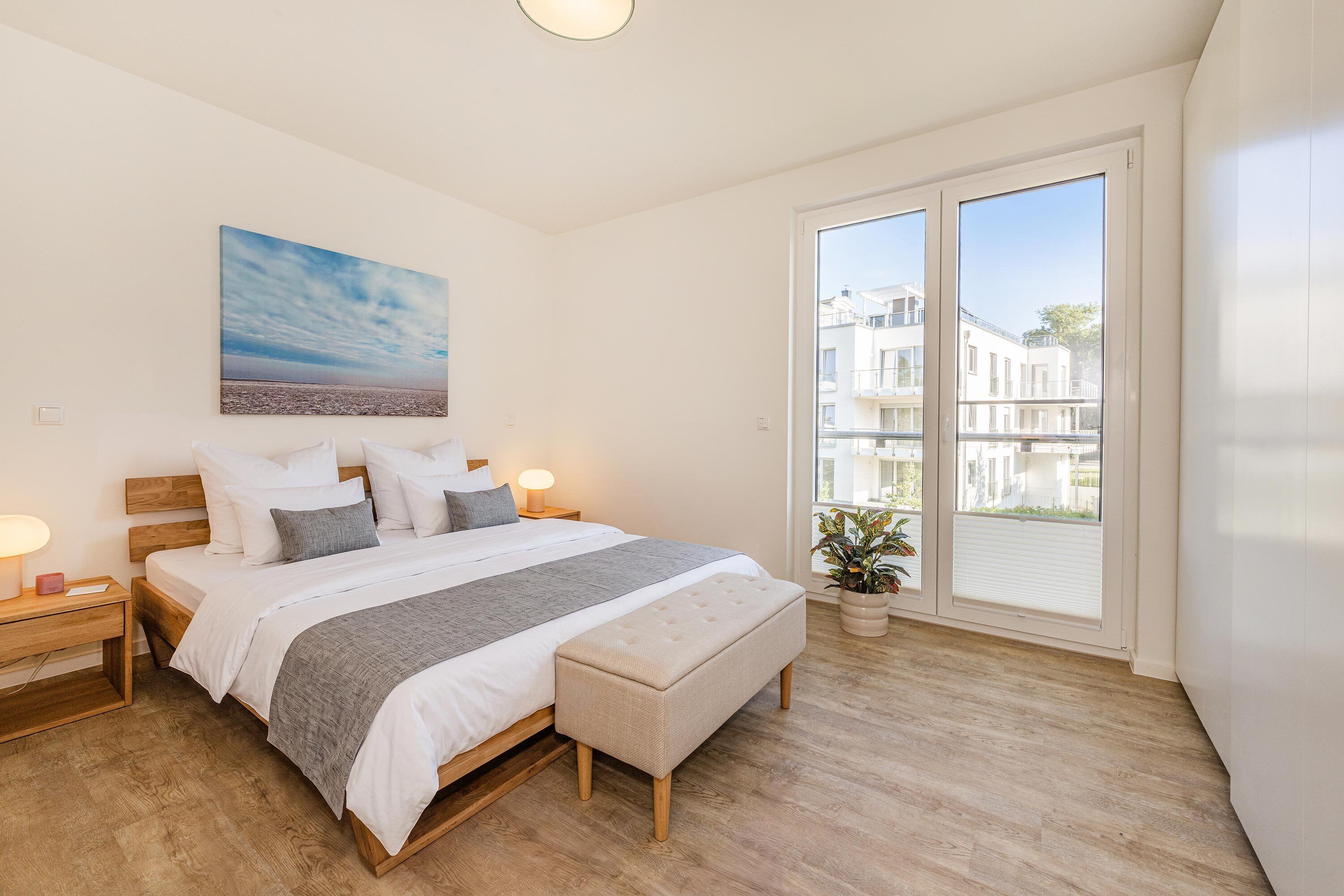 A bedroom with a large bed, bedside lamps, a nightstand, a window with a view of buildings, and a painting on the wall.