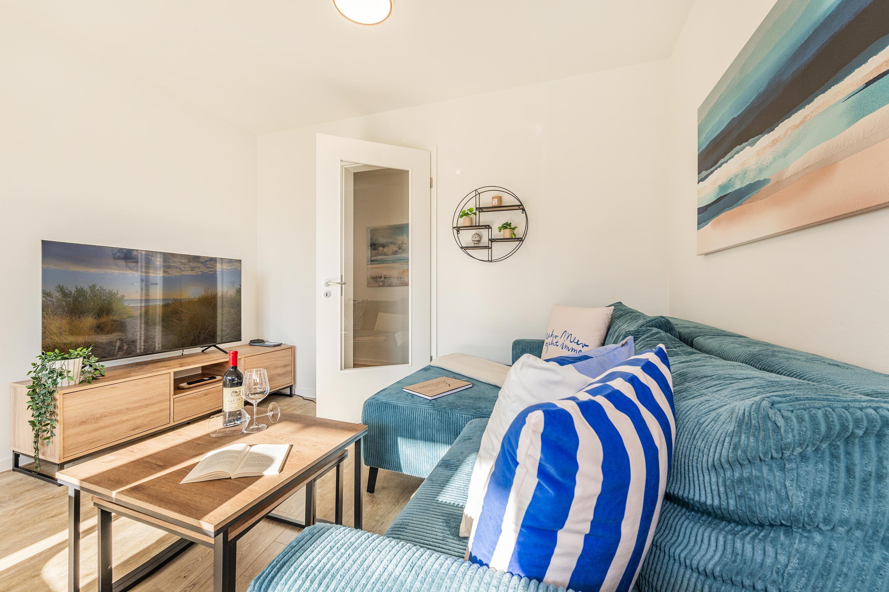 Een moderne woonkamer met een televisie, een houten tafel met een boek, een bank met gestreepte kussens en een schilderij aan de muur.