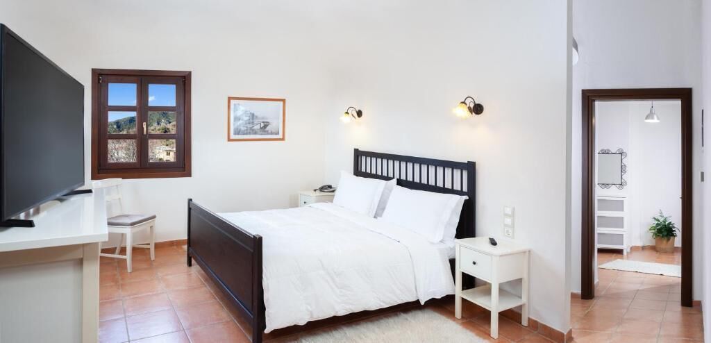 Een slaapkamer met een bed, nachtkastjes, een bureau, een stoel, een televisie, een raam en een ingelijst kunstwerk aan de muur.