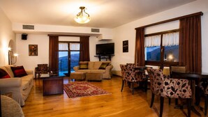 A living room with a sofa, a TV, a dining table with chairs, and a large window with curtains. - Erofili Hotel - Junior Suite with Panoramic View (Portaria)
