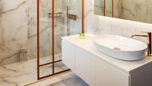 A modern bathroom with a glass-enclosed shower, a white vanity with a gold faucet, and marble walls. - Erofili Hotel, Grand Luxury Suite, Panoramic View (Portaria)