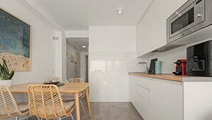 Cozinha moderna com armários brancos, mesa de jantar de madeira com cadeiras de vime, um quadro na parede e vista para o cômodo adjacente.