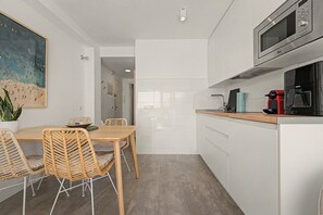 Eine moderne Küche mit weißen Schränken, einem Holzespeisetisch mit Korbstühlen, einem Bild an der Wand und Blick in den angrenzenden Raum.
