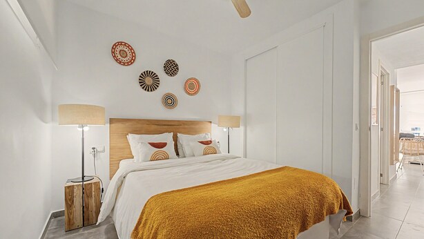 Quarto moderno com cabeceira de madeira, cama grande com roupa de cama branca e um cobertor amarelo mostarda, duas luminárias de mesa e arte decorativa na parede.