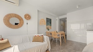 Uma sala de estar moderna com sofá, mesa de jantar e cadeiras. Há um espelho fixado na parede e um lustre.