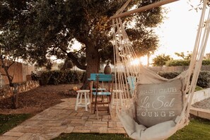 Une chaise suspendue, portant l’étiquette « Beach Club La Sol Lounge », se trouve dans un jardin, près d’un arbre, d’un banc en bois et d’une petite table.