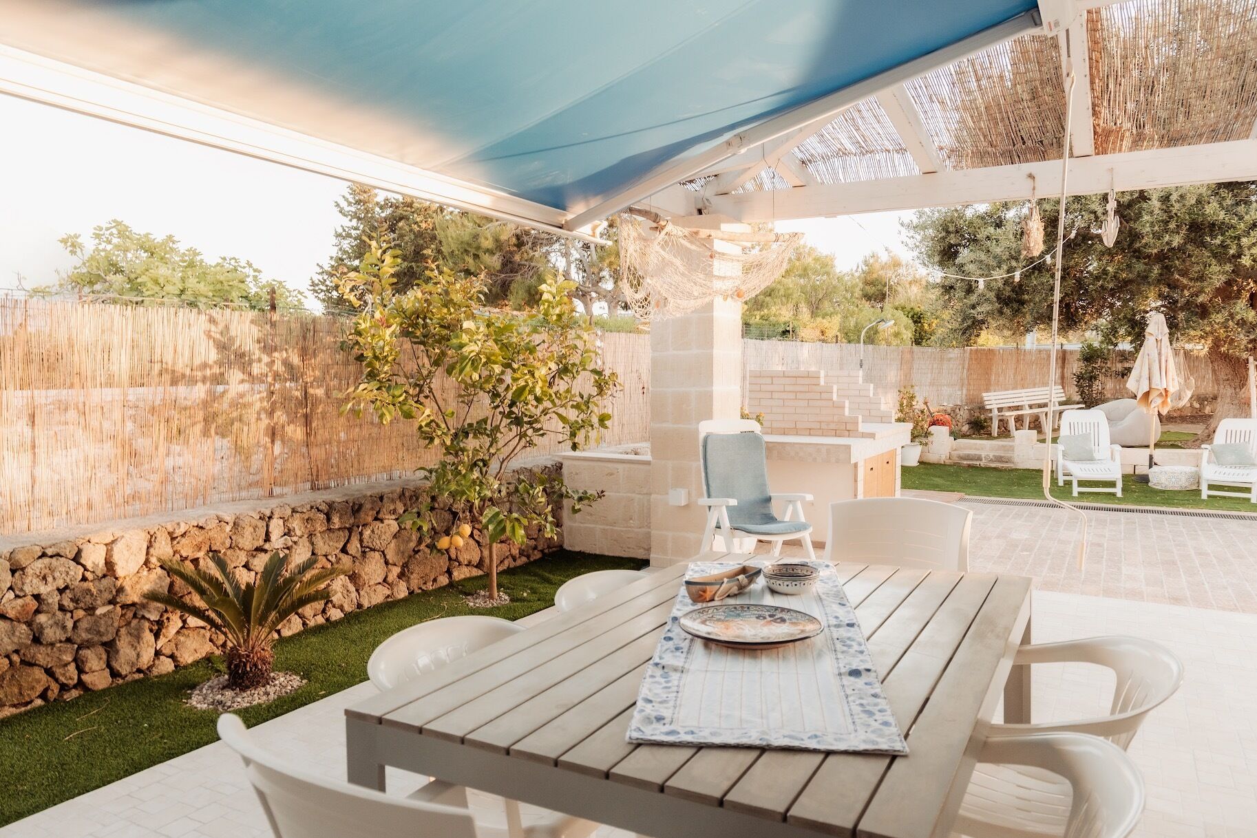Une terrasse extérieure avec une table dressée pour le repas, entourée de chaises blanches, sous une tonnelle bleue.