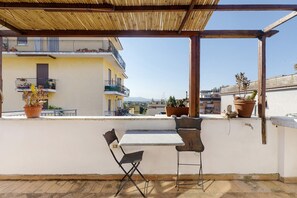 Una terraza en la azotea con mesa y sillas, plantas en macetas y vistas de la ciudad.