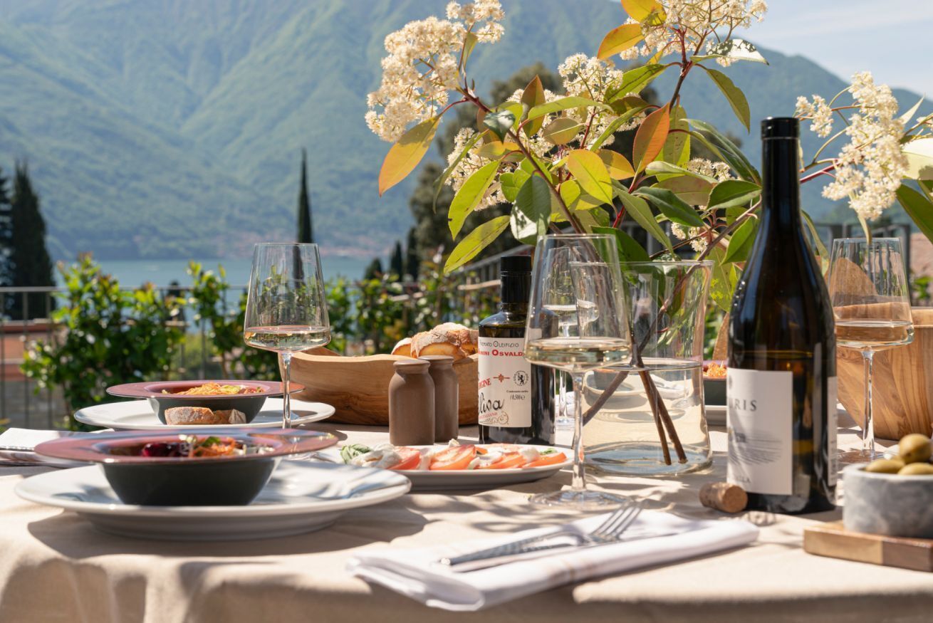 An outdoor dining table with various dishes, wine glasses, and a bottle of wine, set against a mountainous backdrop.