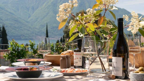 An outdoor dining table with various dishes, wine glasses, and a bottle of wine, set against a mountainous backdrop. - MC Lago - My Home in Como (Tremezzo)