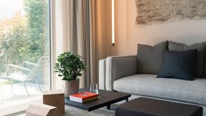 A modern living room with a sofa, a coffee table, and a potted plant. There is a window with curtains and a view of greenery outside. - MC Lago - My Home in Como (Tremezzo)