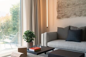 Ein modernes Wohnzimmer mit einer Couch, einem Couchtisch und einem Topf mit Pflanze. Ein Fenster mit Vorhängen bietet Blick auf viel Grün draußen.