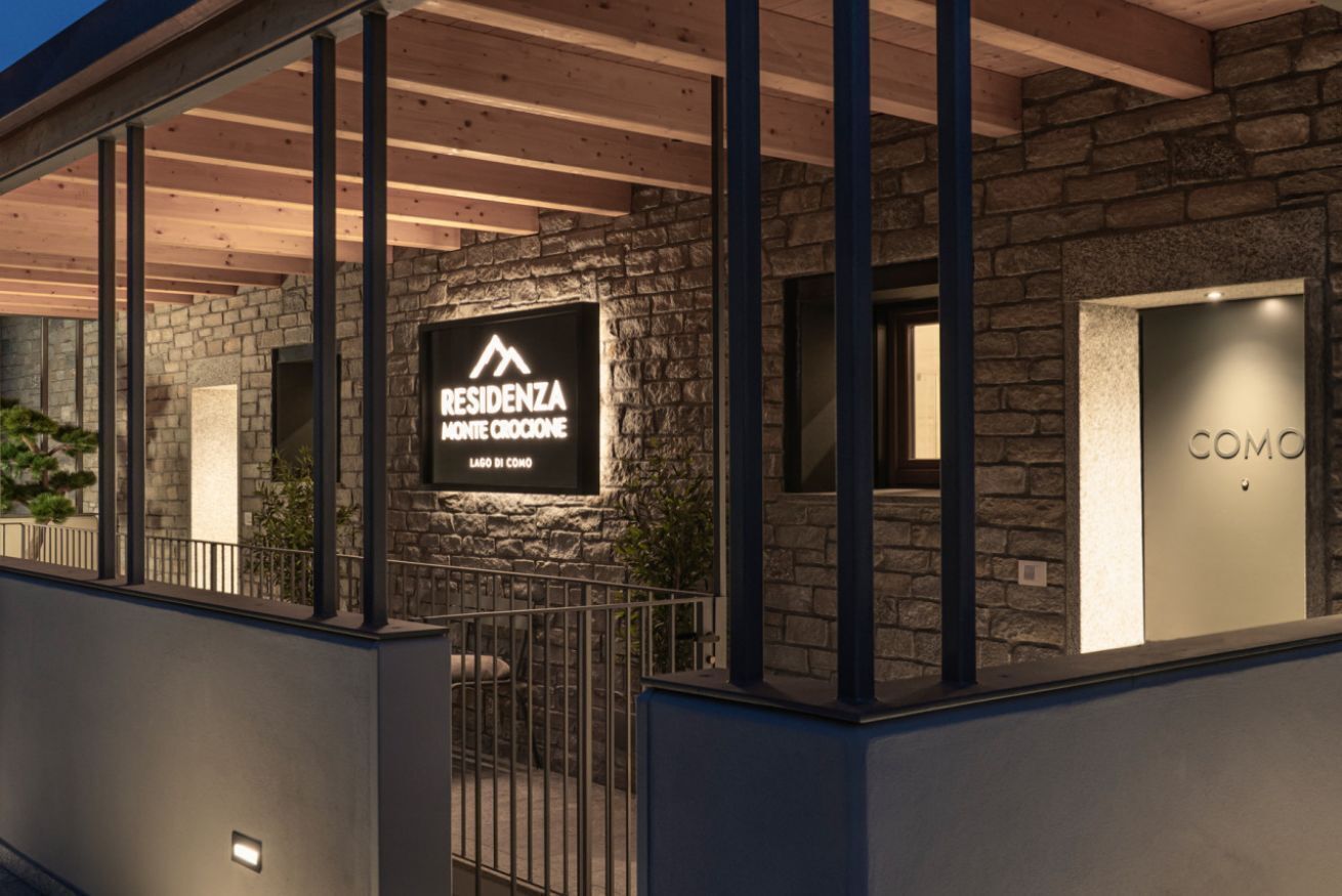 Un bâtiment moderne avec une façade en pierre, une enseigne bien visible indiquant « RESIDENZA MONTI CROCONE LAGO DI CONO », et une porte portant l’inscription « COMO ».