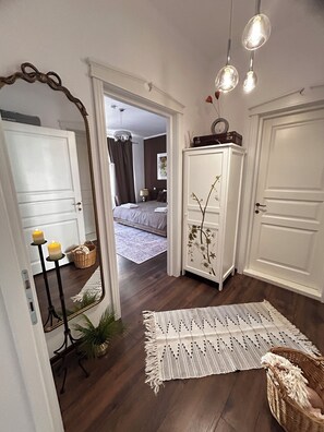 Un couloir avec un miroir, une porte blanche, un meuble blanc orné de motifs floraux, un tapis à motifs et un panier en osier.