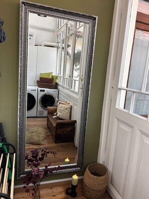 Ein Zimmer mit einem großen Spiegel, der einen Waschbereich, eine hölzerne Bank, einen gewebten Korb und eine Kerze auf dem Boden reflektiert.