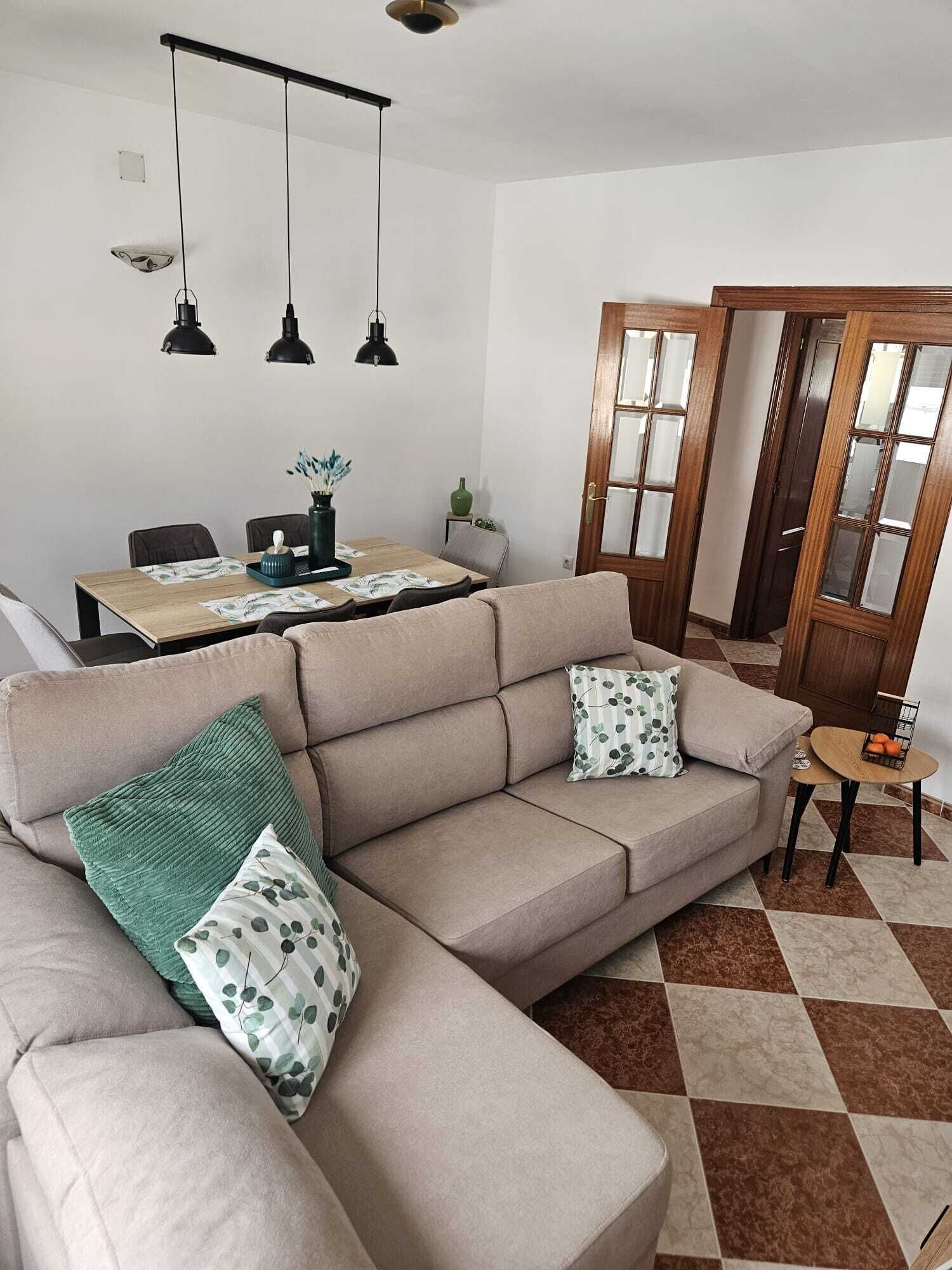 Uma sala de estar com um sofá bege, uma mesa de jantar e uma pequena mesa lateral.