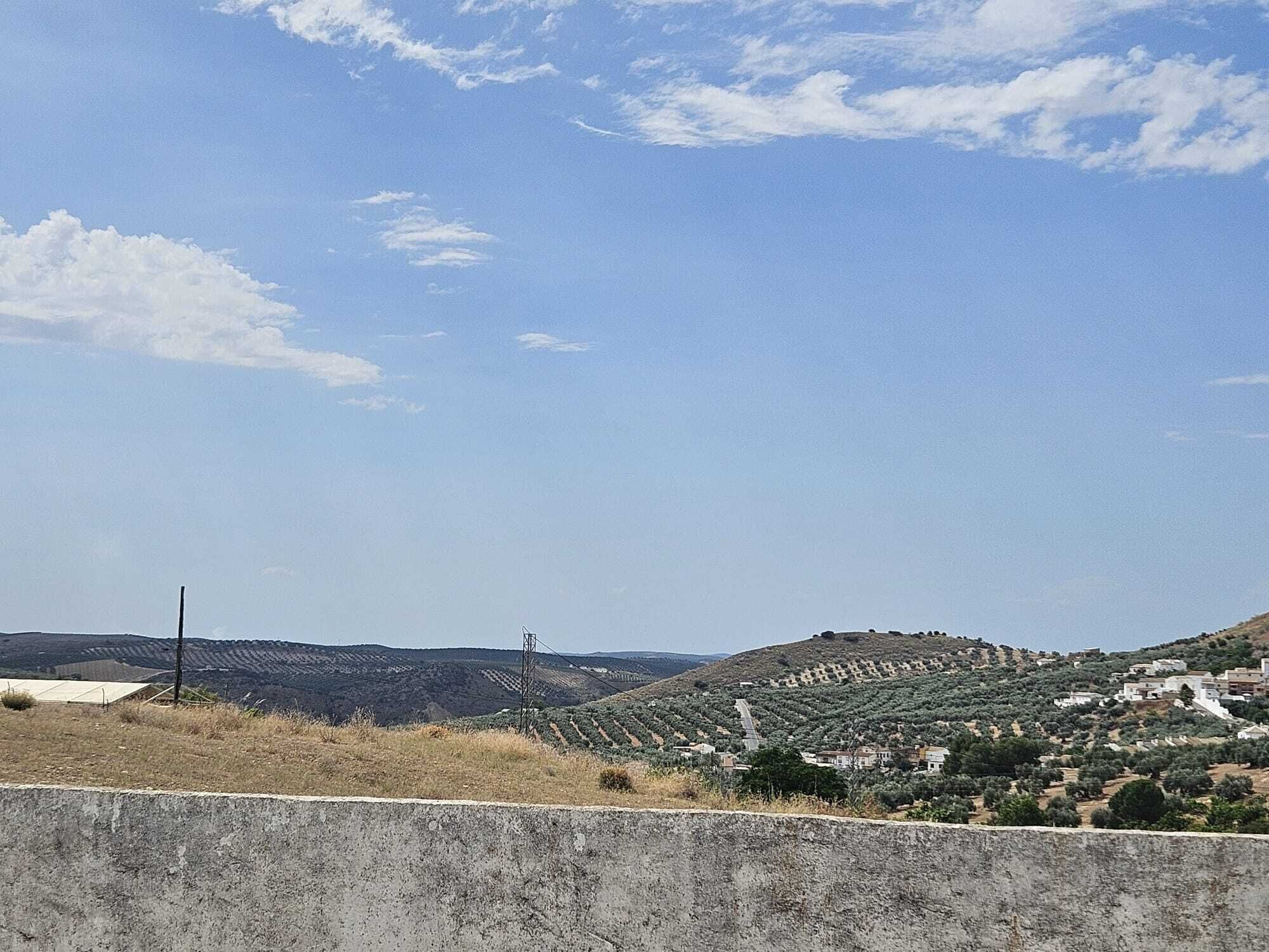 A rural landscape with a clear sky, a concrete barrier in the foreground, and a hilly area with buildings and vegetation.