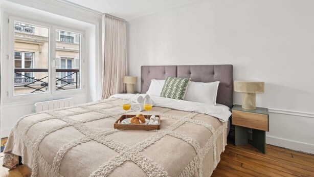 Een net opgemaakt bed met een beige deken, een nachtkastje met een lamp en een raam met gordijnen.