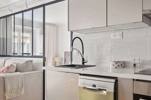 A modern kitchen with a built-in dishwasher, a large window with curtains, and a white countertop. - Charming 1BR near Louvre & Tuileries (Paris)