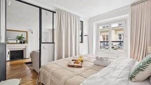 Een slaapkamer met een bed, een nachtkastje, een raam met gordijnen, uitzicht op gebouwen en een open haard in de hoek.