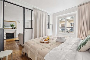 A bedroom with a bed, a bedside table, a window with curtains, a view of buildings outside, and a fireplace in the corner. - Charming 1BR near Louvre & Tuileries (Paris)