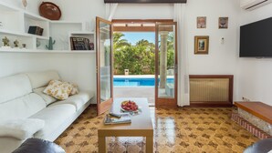 A living room with a sofa, a coffee table with a bowl of fruit, a television, and a view of a swimming pool through an open door. - Villa 'Russadir' with Private Pool, Wi-Fi and Air Conditioning (Santanyí)