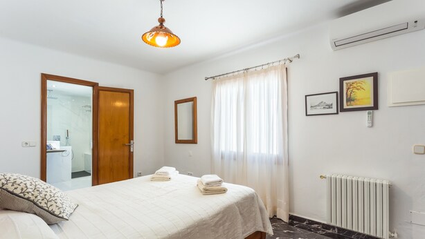 A hotel room with a bed, a bathroom visible through an open door, a radiator, and a window with curtains. - Villa 'Russadir' with Private Pool, Wi-Fi and Air Conditioning (Santanyí)