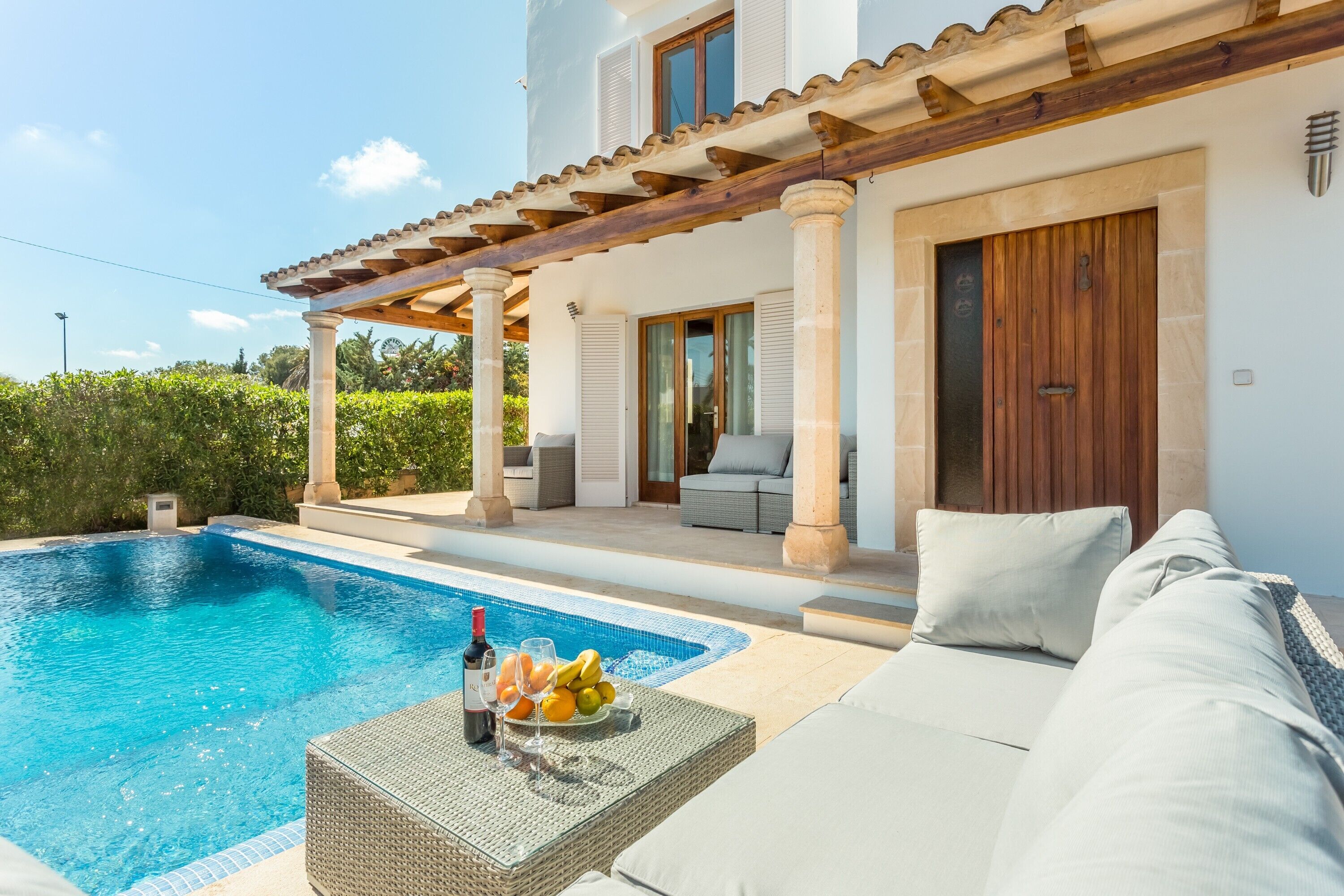 A poolside area with a lounge chair, a wicker table with a bottle and glasses, and a fruit bowl.