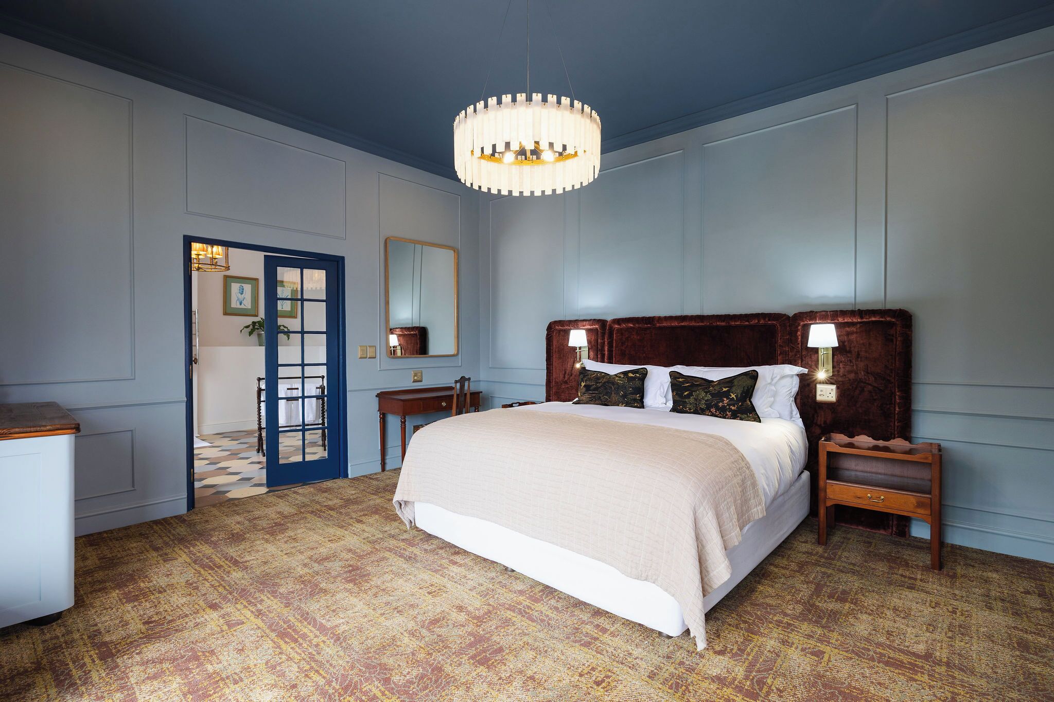 Une chambre d’hôtel avec un grand lit, un lustre, une porte bleue et une moquette à motifs.