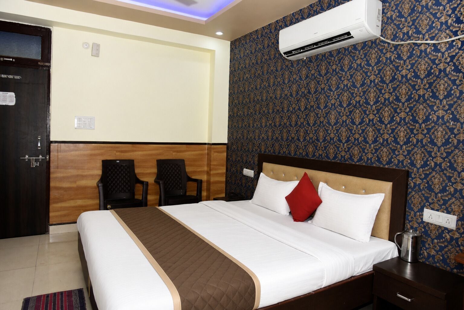 Habitación de hotel con cama, dos sillas, equipo de aire acondicionado, mesita de noche y pared con diseño.