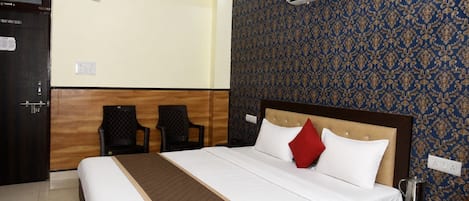 A hotel room with a bed, two chairs, an air conditioning unit, a nightstand, and a patterned wall.